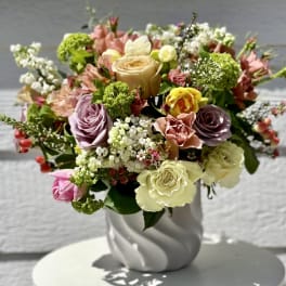 Mixed arrangement of cream, peach, lavender, and yellow roses with small blooms in a white ceramic vase