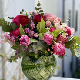 Bright pink and red tulips, roses, and ranunculus arranged in a round green glass vase