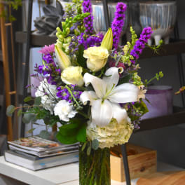 Tall arrangement of white lilies and roses with purple flowers in a green glass vase