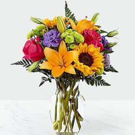 Colorful bouquet with sunflowers, lilies, roses, and purple blooms in a clear glass vase