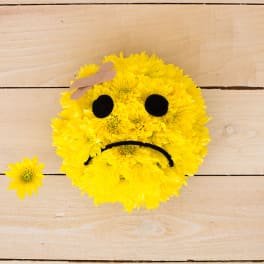 Round yellow chrysanthemum arrangement shaped like a sad face with bandage on forehead on wooden surface