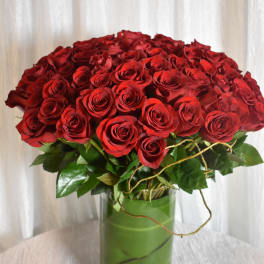 Large arrangement of red roses in a clear cylinder vase wrapped with green leaves.