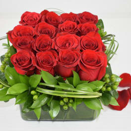 Square arrangement of red roses with decorative green loops in a glass cube vase