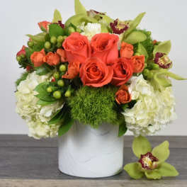 Low arrangement of orange roses, green orchids, and white hydrangeas in a white vase with one orchid bloom beside it.