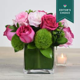 Short arrangement of pink roses and soft pink blooms in a green-lined glass cube vase.