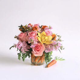 Short arrangement of pink roses, yellow orchid, and orange flowers in a carrot-filled glass vase with a carrot beside it