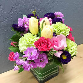 Colorful bouquet in a square glass vase with tulips, carnations, and purple blooms