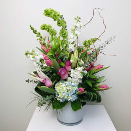 Tall arrangement of pink roses and tulips with green Bells of Ireland, white lilies, and blue hydrangea in a white vase