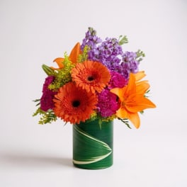 Orange gerberas and lilies in a green vase with purple blooms