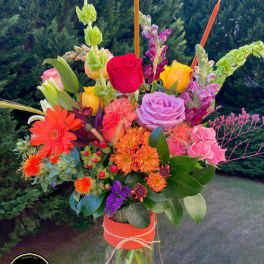 Tall mixed bouquet of bright roses, gerbera daisies, and mums in a glass vase wrapped with orange ribbon.