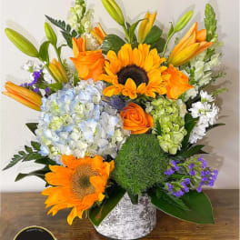Bright arrangement of sunflowers, orange roses, lilies and hydrangeas in a white cylinder vase