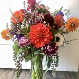 Tall mixed bouquet of orange, red, purple, and white flowers arranged in a clear glass vase.