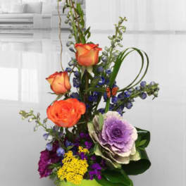 Tall mixed flower arrangement in a lime green vase with orange roses and purple blooms