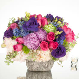 Low arrangement of pink roses, purple blooms, and pale hydrangeas in a silver vase