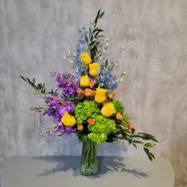 Tall mixed bouquet in a clear glass vase with yellow, purple, blue, and green blooms