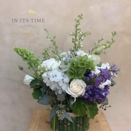 Mixed bouquet of white, green, and purple flowers in a bamboo-wrapped container