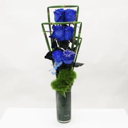 Tall modern arrangement of vivid blue roses in a green-lined glass cylinder with bamboo accents and fuzzy green dianthus