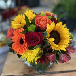 Mixed bouquet of roses, sunflowers, and gerbera daisies in a glass vase