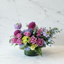 Low arrangement of mauve roses with purple mums and green and blue hydrangea in a glass vase