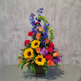 Tall mixed flower arrangement with sunflowers, roses, and blue delphinium in a dark vase
