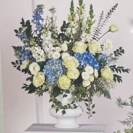 Large floral arrangement in a white pedestal vase with blue, white, and pale yellow blooms