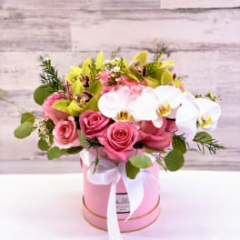 Pink roses and white orchids arranged in a pink hat box with a ribbon