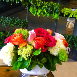 Low arrangement of red, orange, and pink roses with white hydrangeas in a white bowl vase