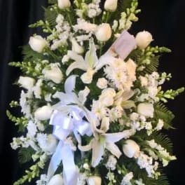 White floral standing spray with lilies, roses, and ribbon on a dark backdrop