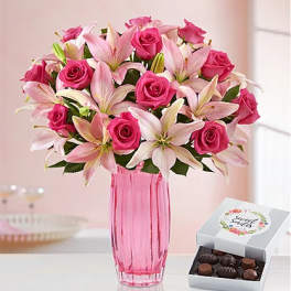 Pink roses and lilies in a pink glass vase beside a box of chocolates