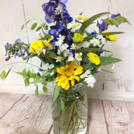 Wildflower-style bouquet of blue delphinium, yellow daisies, and white blooms in a clear glass jar vase