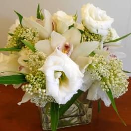 Low arrangement of white roses, lisianthus, and orchids in a clear glass cube vase