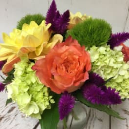 Low arrangement of orange rose, green hydrangea, yellow blooms and purple flowers in a clear glass vase