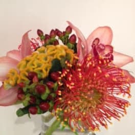 Compact arrangement of pink lilies, red pincushion protea, yellow blooms, and berries in a clear glass vase.