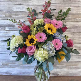 Mixed bouquet of pink roses, gerbera daisies, sunflowers, and hydrangeas in a glass vase