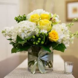 Yellow roses and white flowers arranged in a clear glass vase with a ribbon.
