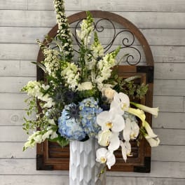 Tall floral arrangement with white orchids, blue hydrangeas, and pale blooms in a white vase