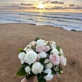 Bridal bouquet of white and blush roses and ranunculus held by the ocean at sunset