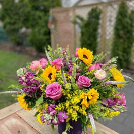 Mixed bouquet of pink roses, yellow sunflowers, and purple flowers in a purple vase