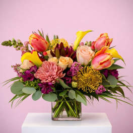 Colorful arrangement of yellow calla lilies, roses, tulips, and protea in a clear glass cube vase