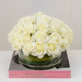 White roses arranged in a low glass bowl on a pink book box.