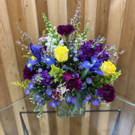 Mixed bouquet of yellow roses, blue irises, and purple flowers in a glass vase