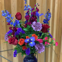 Tall mixed arrangement with red roses, purple delphinium and carnations in a blue glass vase