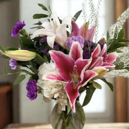 Pink lilies, white hydrangea, and purple flowers arranged in a clear glass vase on a wooden table