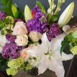Mixed arrangement of purple lilacs, white lilies, and pale roses in a clear glass vase