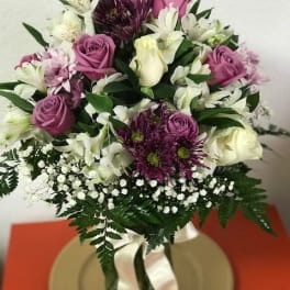 Round arrangement of purple and white flowers in a clear vase with white ribbon on a gold plate