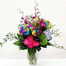 Tall arrangement of rainbow roses, orchids, and bright mixed blooms in a clear glass vase