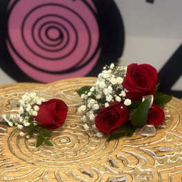 Two red rose boutonnieres with white filler flowers resting on a carved wooden surface