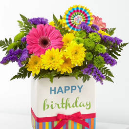 Bright birthday bouquet with pink gerbera, yellow blooms, and purple accents in a happy birthday cube vase