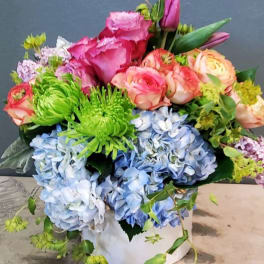 Colorful arrangement of pink roses, green mums, and blue hydrangeas in a white cylinder vase.