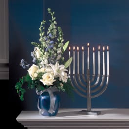 White roses and lilies in a blue vase beside a lit menorah
